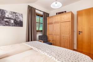 Un dormitorio con cama, un armario de madera, una silla, una ventana con cortinas y un cuadro en la pared.