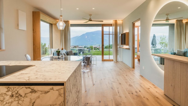 Uma cozinha moderna com uma ilha de mármore, uma área de refeições com cadeiras transparentes e vista para montanhas através de grandes janelas.