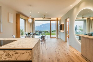 Una cocina moderna con una isla de mármol, un comedor con sillas transparentes y vistas a montañas a través de grandes ventanales.