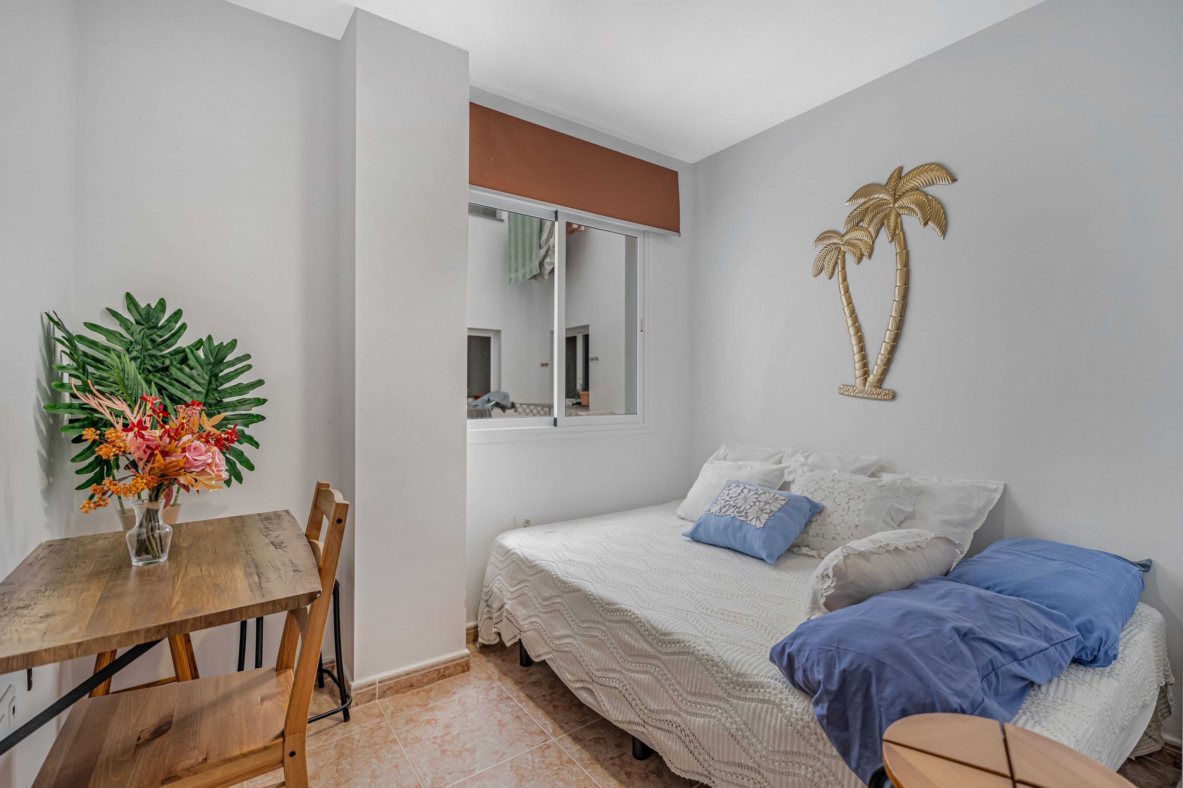 Ein Schlafzimmer mit einem Bett, einem kleinen Tisch mit Blumenvase, einem Fenster und einer Wanddekoration.