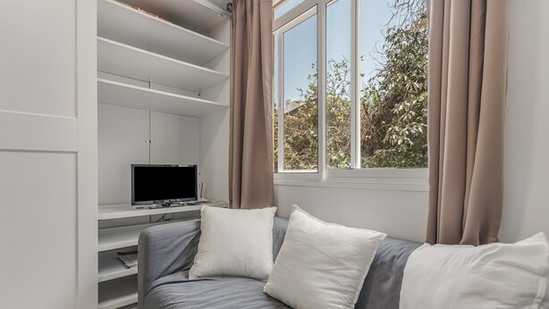 A modern living room with a grey sofa, white cushions, a flat-screen TV, and a window with curtains.