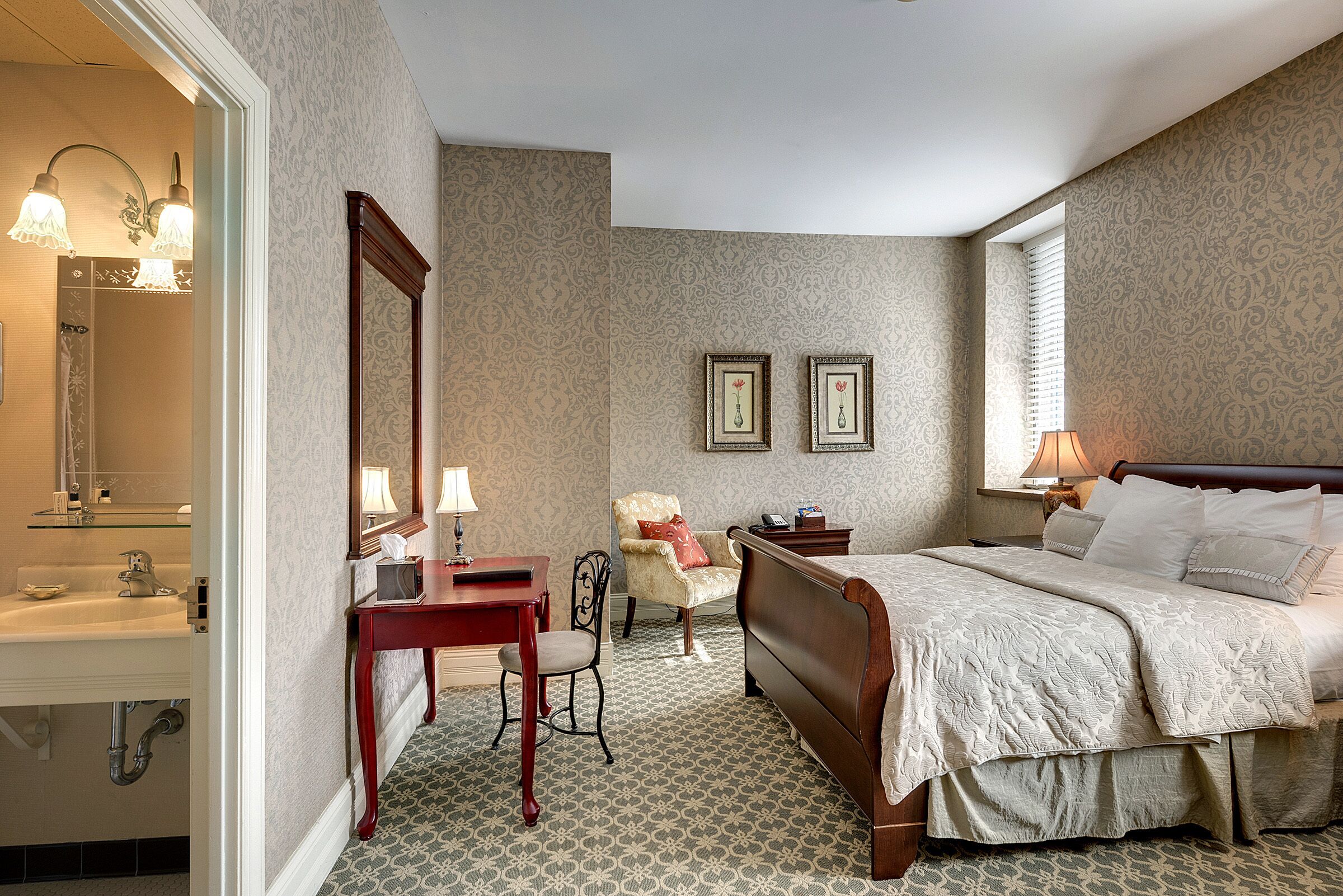 A hotel room with a bed, a desk with a lamp, a chair, a mirror, and a window.