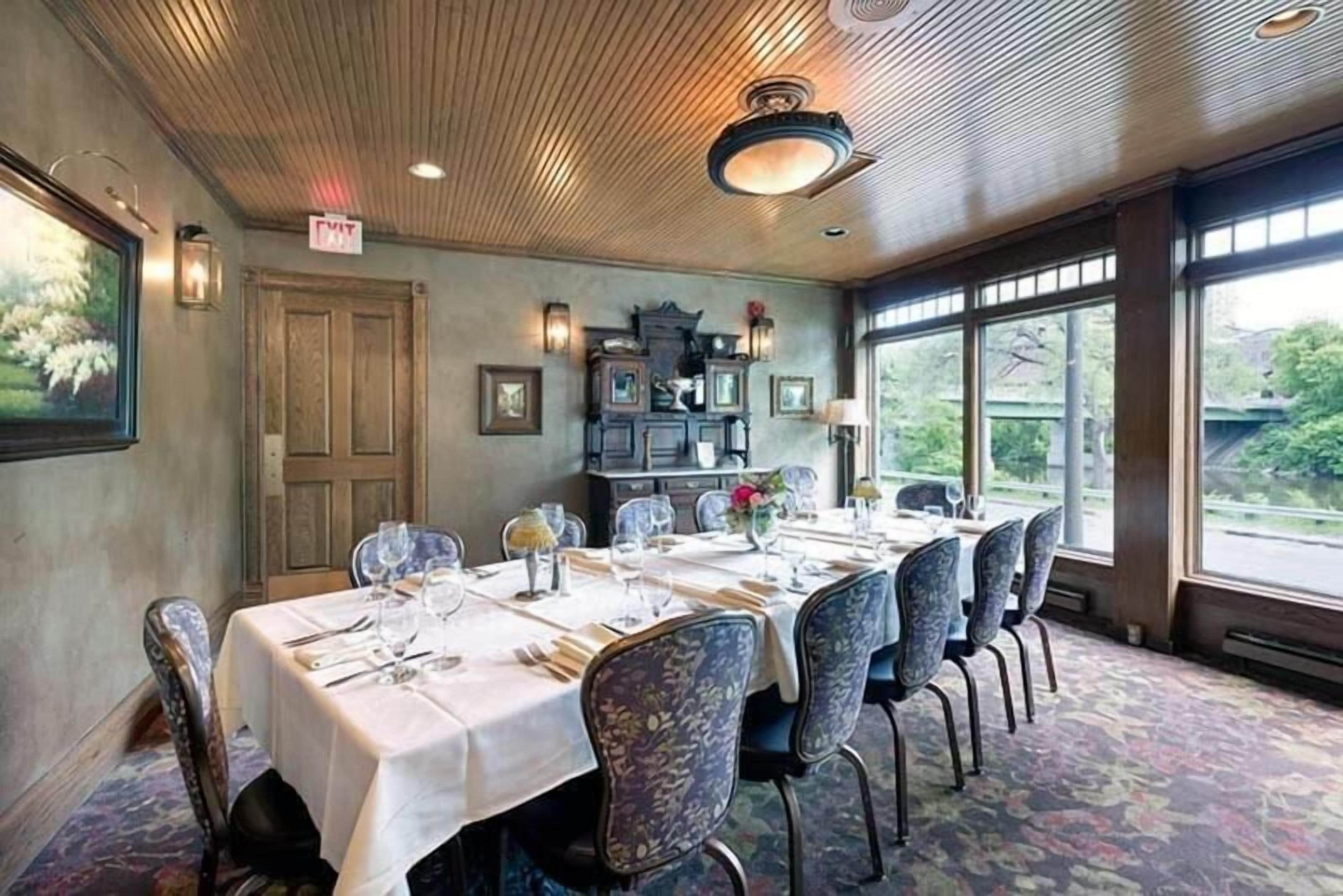A dining room with a long table set for six, flanked by chairs with floral upholstery. A wooden ceiling with a central light fixture is visible, and a large window offers a view of greenery outside.