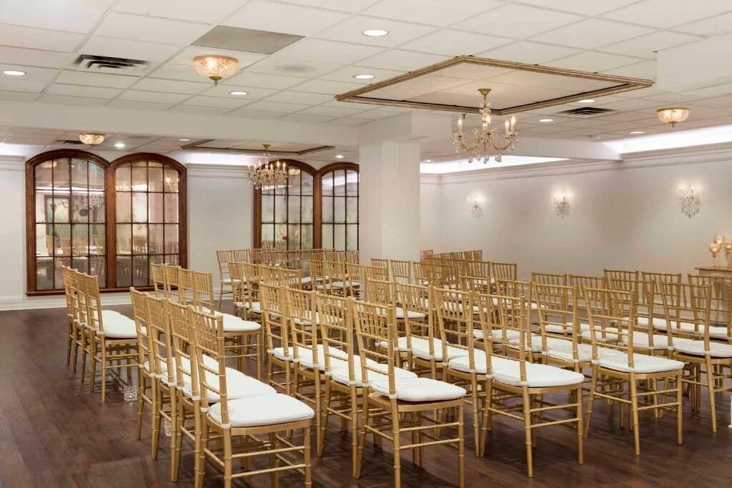 Ein geräumiger Raum mit Reihen goldener Stühle und weißen Kissen, großen Fenstern mit Holzrahmen und eleganten Kronleuchtern.