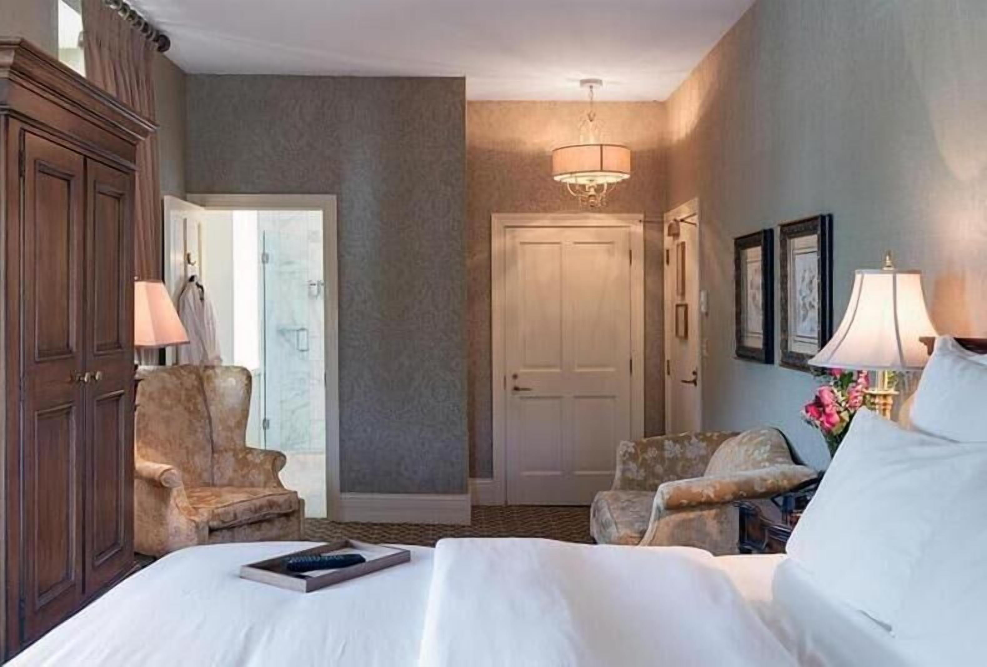 Une chambre d’hôtel avec un grand lit, deux fauteuils, une armoire en bois, une porte et un lustre.
