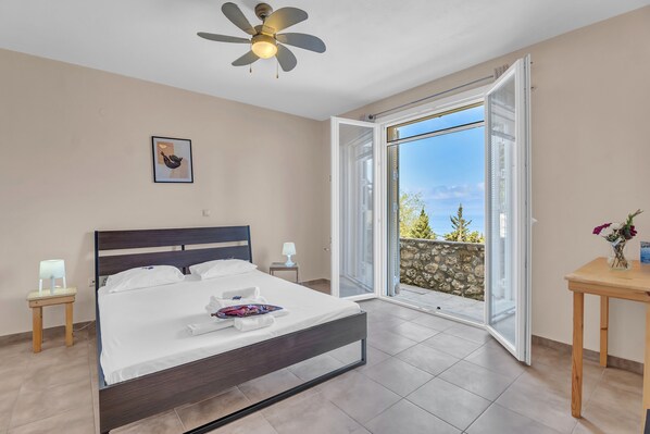 Une chambre avec un lit, un ventilateur de plafond, une table de chevet, une lampe, une table ornée de fleurs et un balcon avec vue.