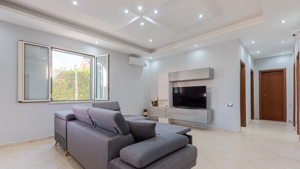 A modern living room with a grey sectional sofa, a flat-screen TV mounted on a grey wall, and a large window providing natural light. - Villa 'Villino Giovanna' with Private Terrace, Wi-Fi and Air Conditioning (Trabia)