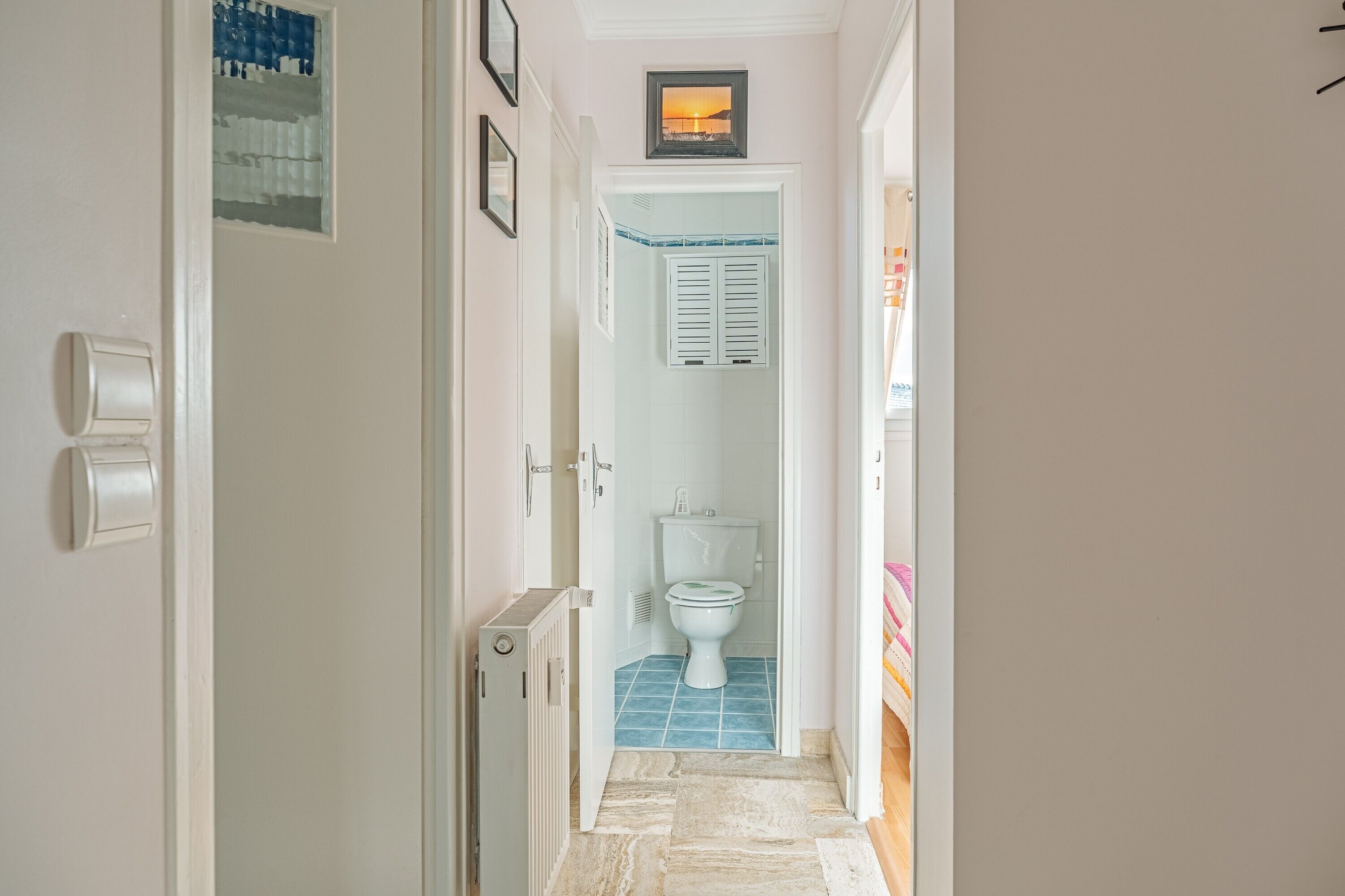 Ein schmaler Flur, der zu einem Badezimmer mit Toilette, Heizkörper und Fliesenboden führt.