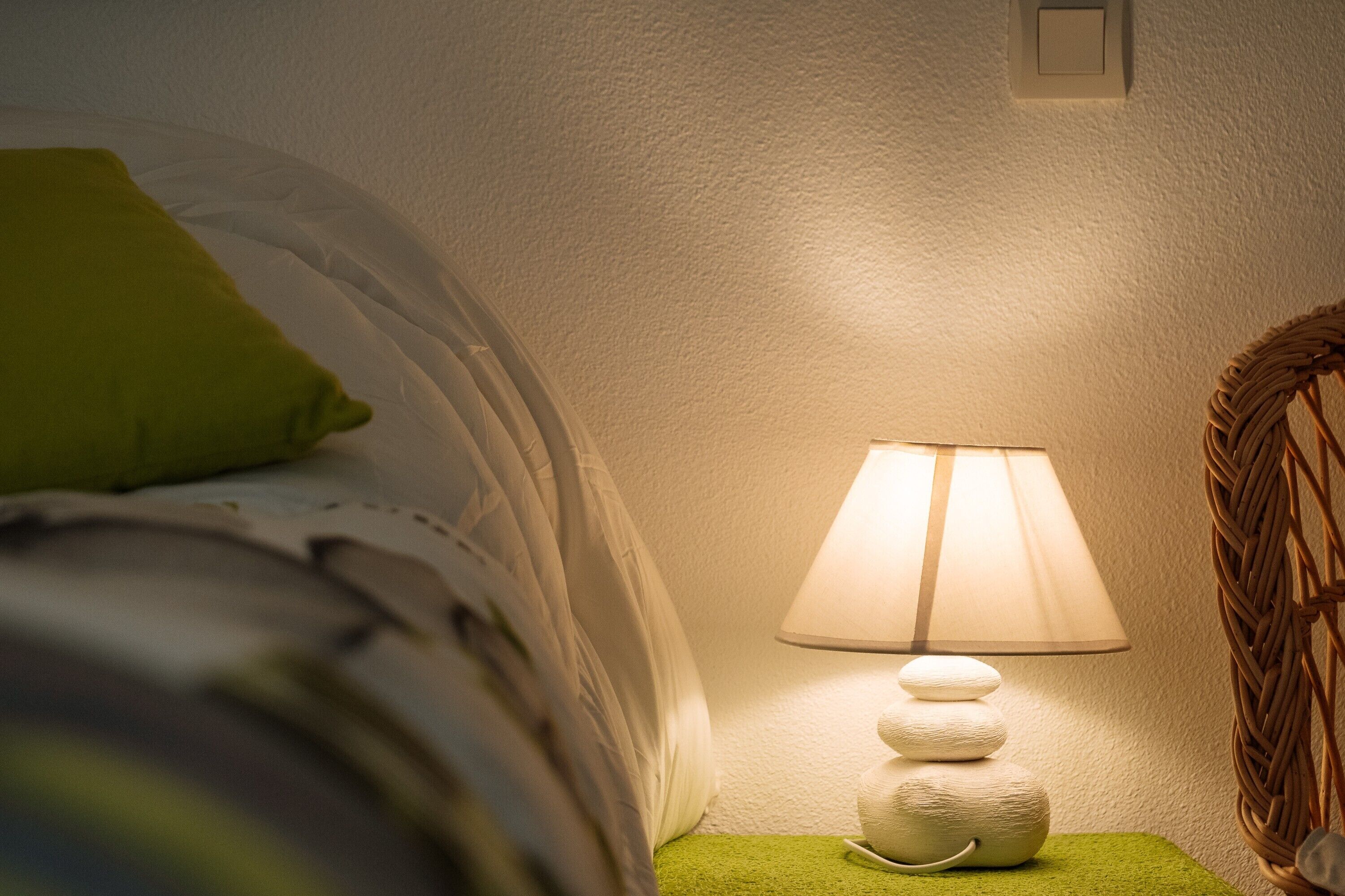 En seng med hvidt sengetøj og en grøn pude, en sengebordslampe med hvid skærm, og en vævet kurv på en grøn pude.