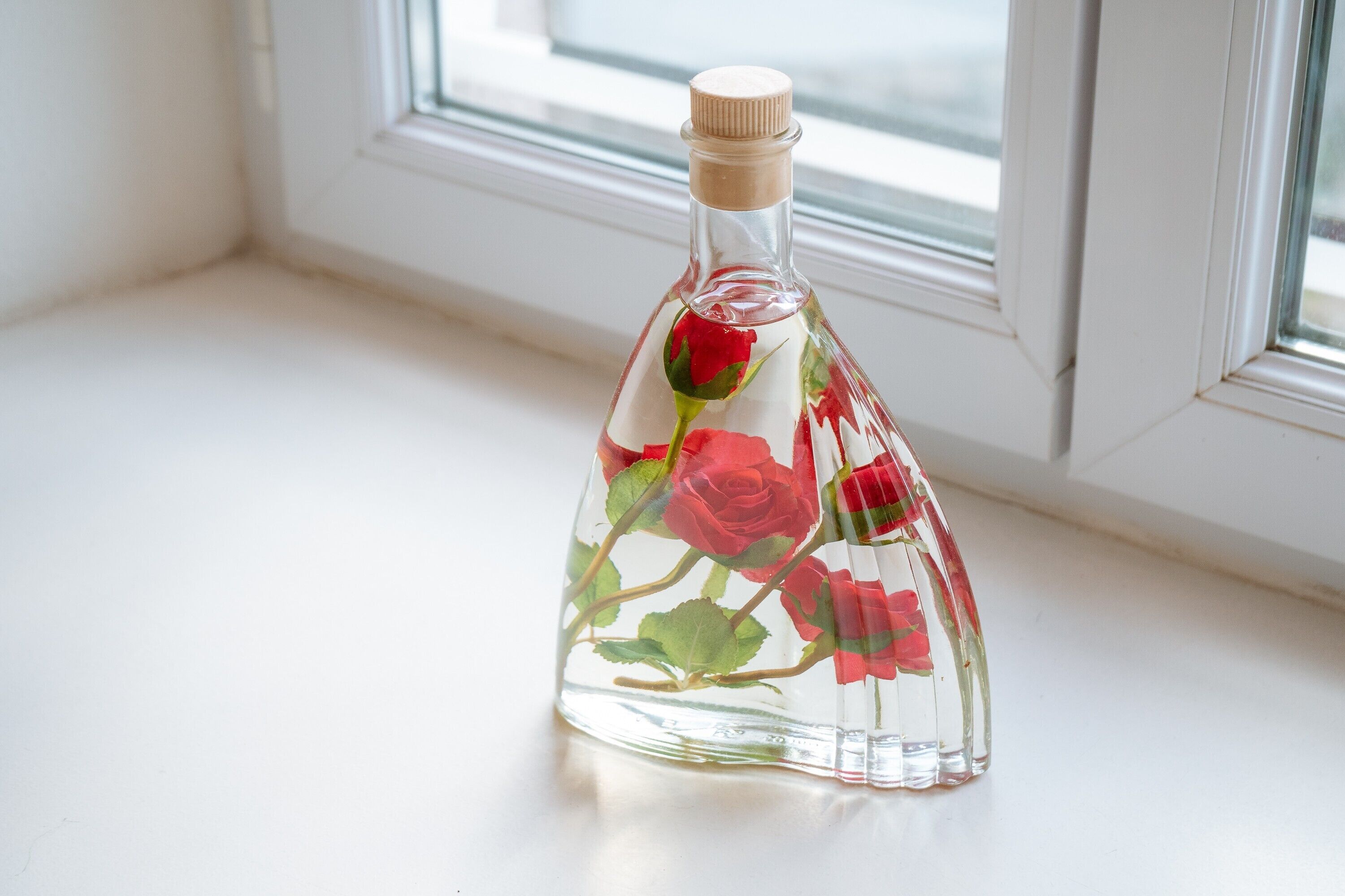 Een transparante glazen fles met een bloemendecor op een wit oppervlak, in de buurt van een raam.