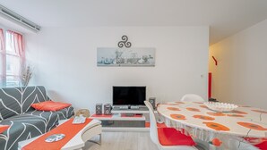 A modern living room with a sofa, TV, speakers, and a dining table with red cushions.