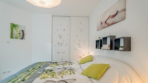 A bedroom with a bed, pillows, a wall-mounted shelf with books, a door with a floral decal, and a painting on the wall.
