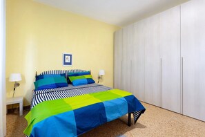 A bed with a striped and patchwork bedspread, two bedside lamps, a framed picture on the wall, and a wardrobe.