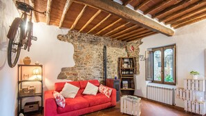 A cozy living room with a red sofa, stone walls, wooden beams, a bookshelf, and a window with a view. - Holiday Home 'L'arco Della Fonte' with Private Terrace, Wi-Fi and Air Conditioning (Gaiole in Chianti)