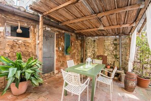 A rustic outdoor dining area with a green table and white chairs, surrounded by stone walls and potted plants.