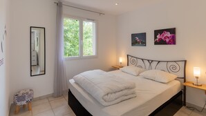 A neatly made bed with a headboard, a bedside table with a lamp, a window with curtains, and a small stool. - Holiday Home 'Le Pequelet' with Shared Pool, Wi-Fi and Air Conditioning (Payzac)
