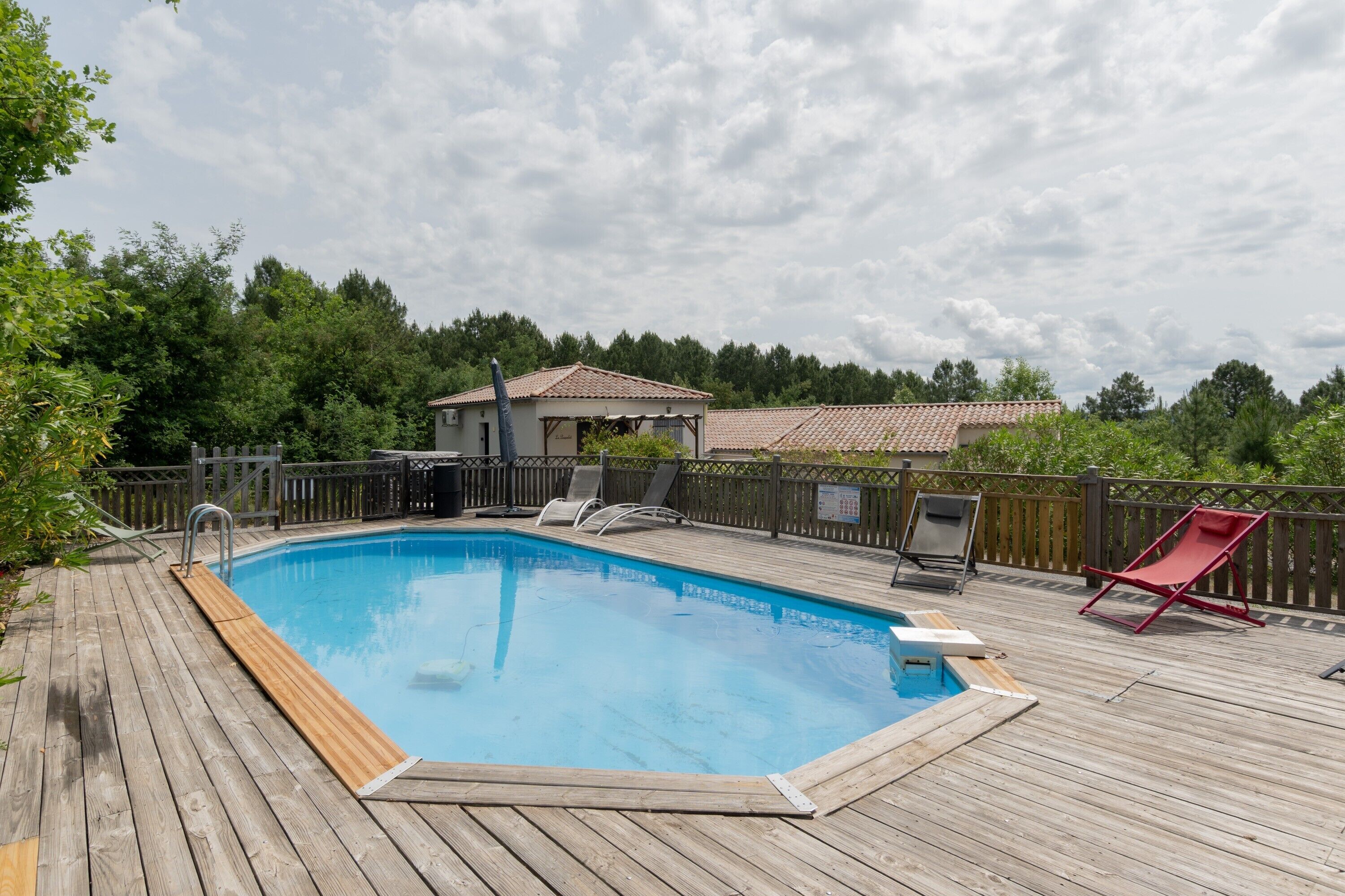 A wooden deck with a swimming pool, lounge chairs, and a small building with a tiled roof.