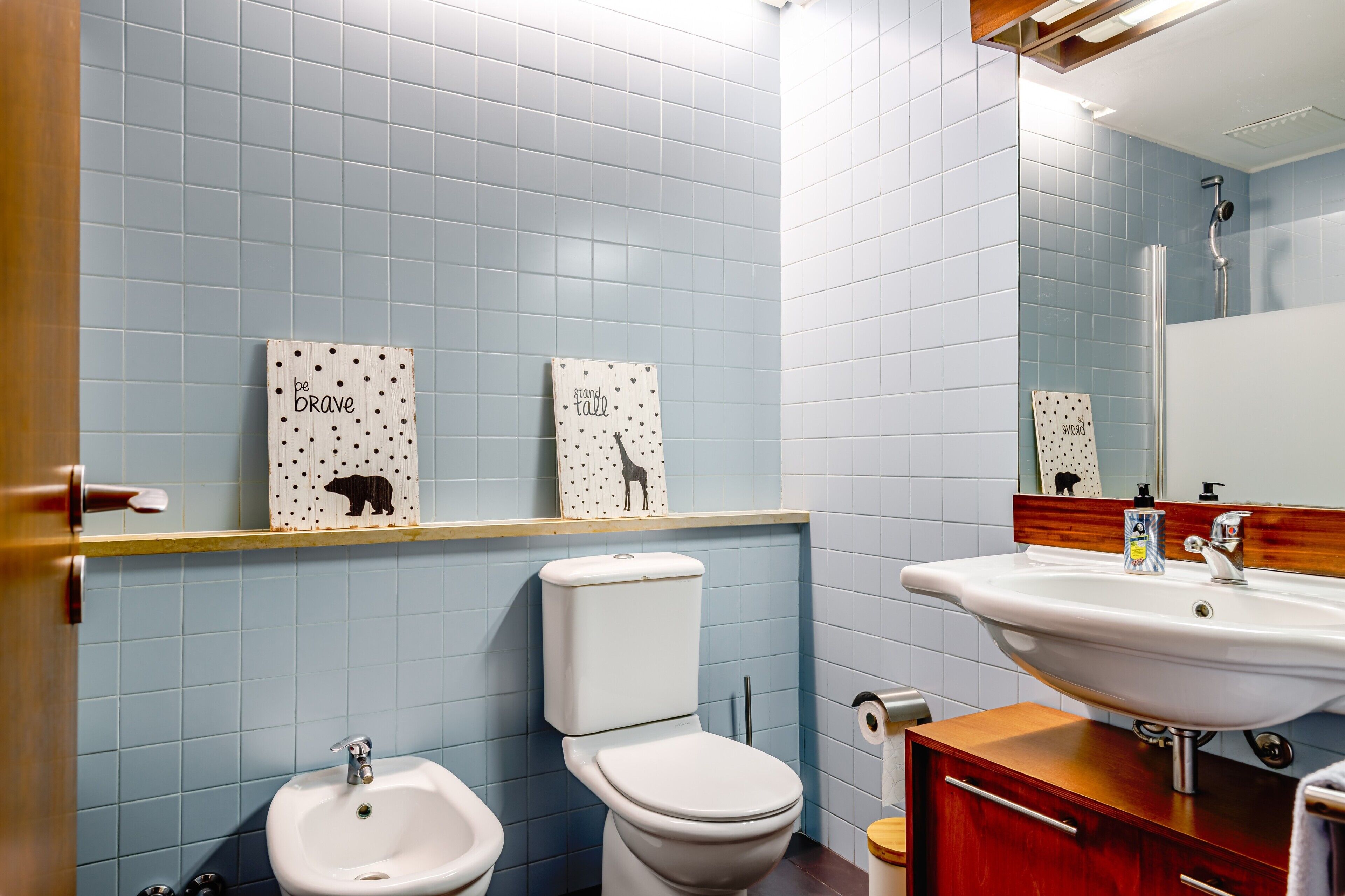 Et badeværelse med toilet, bidet og vask, blå flisevægge og motiverende billedkunst på væggen.