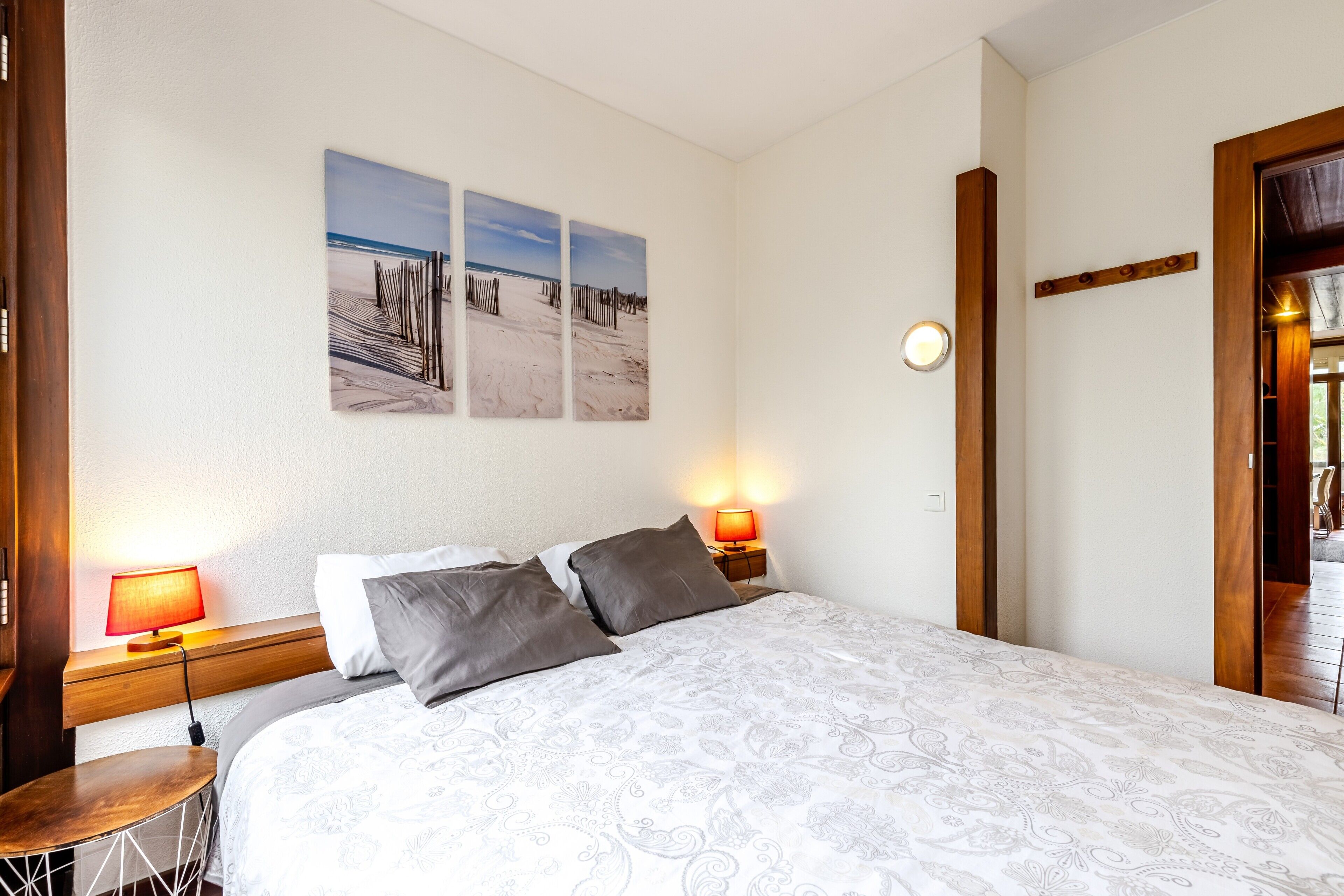 Quarto com cama, duas luminárias de apoio, uma mesa redonda e três obras de arte com tema praiano.