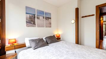 Quarto com cama, duas luminárias de apoio, uma mesa redonda e três obras de arte com tema praiano.