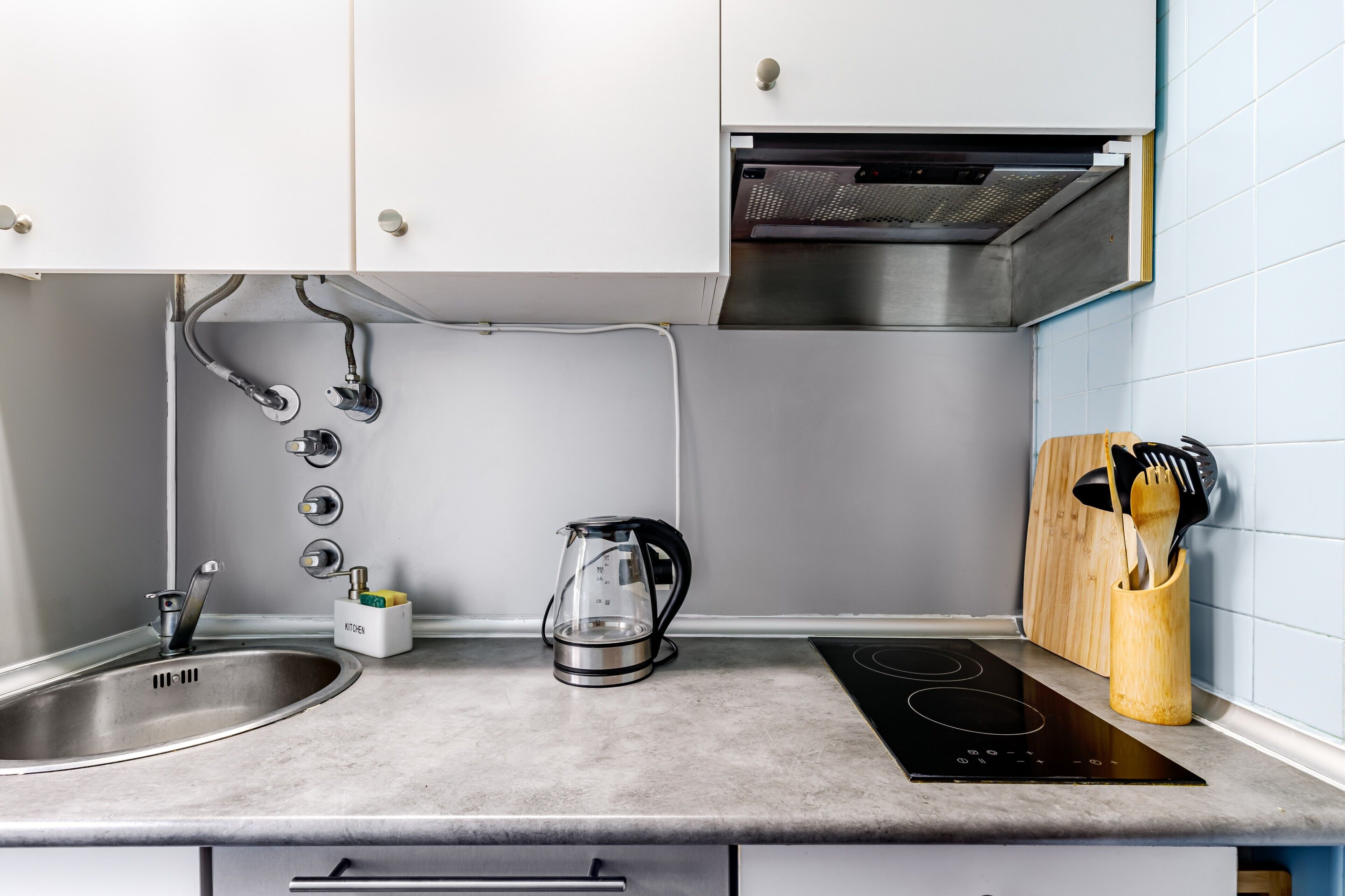 Una cocina con un lavamanos de acero inoxidable, una cafetera de vidrio, una cocina por inducción y utensilios de madera en un soporte.