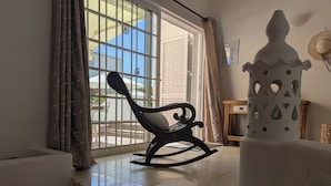 Een kamer met een schommelstoel, een wit decoratief object en uitzicht door een raam met gordijnen.