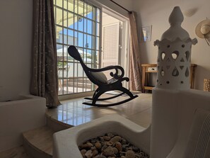 Een kamer met een schommelstoel, een wit decoratief object en uitzicht door een raam met gordijnen.