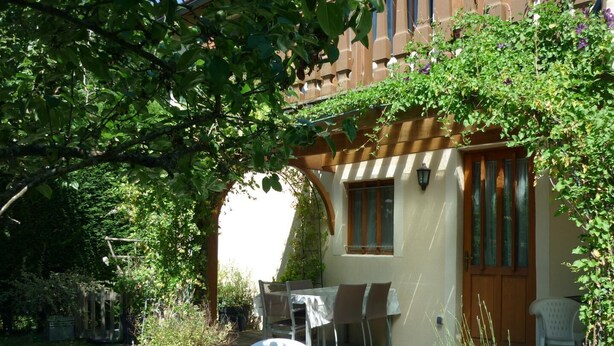 A patio with a table and chairs, surrounded by greenery and a building with a wooden door and windows. - Gîte des Cèdres - Ameugny (AMEUGNY)