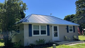 A single-story house with a blue metal roof, a front porch with two chairs, and a small American flag. - Welcome to The Driftwood Cottage! A cozy 2-bedroom home in Grand Marais, MI. (Grand Marais)