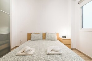 A minimalist hotel room with a bed, two pillows, a nightstand, and a window.