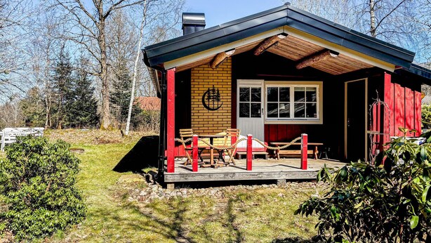 Una cabaña pequeña con una terraza de madera, una puerta roja y una ventana.