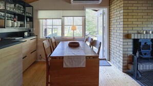 Un comedor con una larga mesa de madera, sillas y una ventana con persianas. Hay una chimenea de ladrillo y una zona de cocina con estantes y utensilios a la vista.
