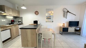 A modern kitchen with a central island, a dining area with chairs, a TV, and a wall-mounted clock. - L'appartement Pins D97 (CAPBRETON)