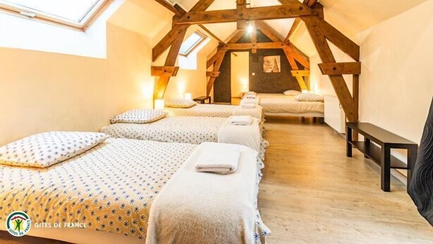 Un dormitorio acogedor en el ático, con techo inclinado, vigas de madera y un tragaluz. Hay tres camas con ropa de cama a juego y un banco en la esquina.