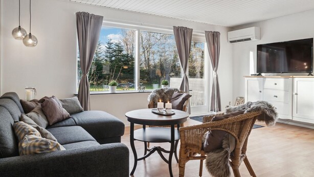Ein modernes Wohnzimmer mit einer Couch, einem runden Esstisch, Korbstühlen, einem Fernseher und Blick auf Bäume durch große Fenster.