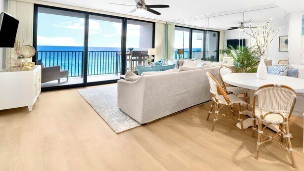 Een moderne woonkamer met een bank, twee rieten stoelen, een eettafel en een groot raam met uitzicht op de oceaan.