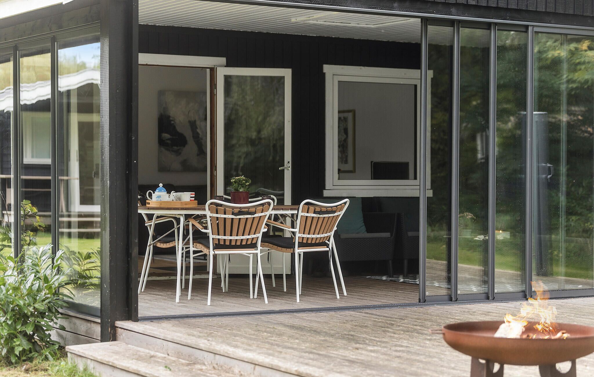 A modern outdoor dining area with a glass enclosure, wooden deck, and a fire pit.