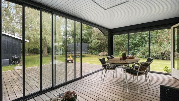 A covered patio with a round table and chairs, large windows, and a view of a backyard with a barbecue grill and trees. - Cozy home in Gilleleje with kitchen (Gilleleje)