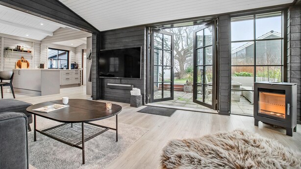 Ein modernes Wohnzimmer mit Kamin, einer Küche im Hintergrund und einer großen Glasdoor, die auf eine Terrasse führt.