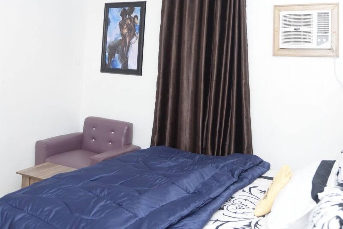 Un dormitorio con cama, una silla, ventilador de techo, ventana con cortinas y aire acondicionado de pared.