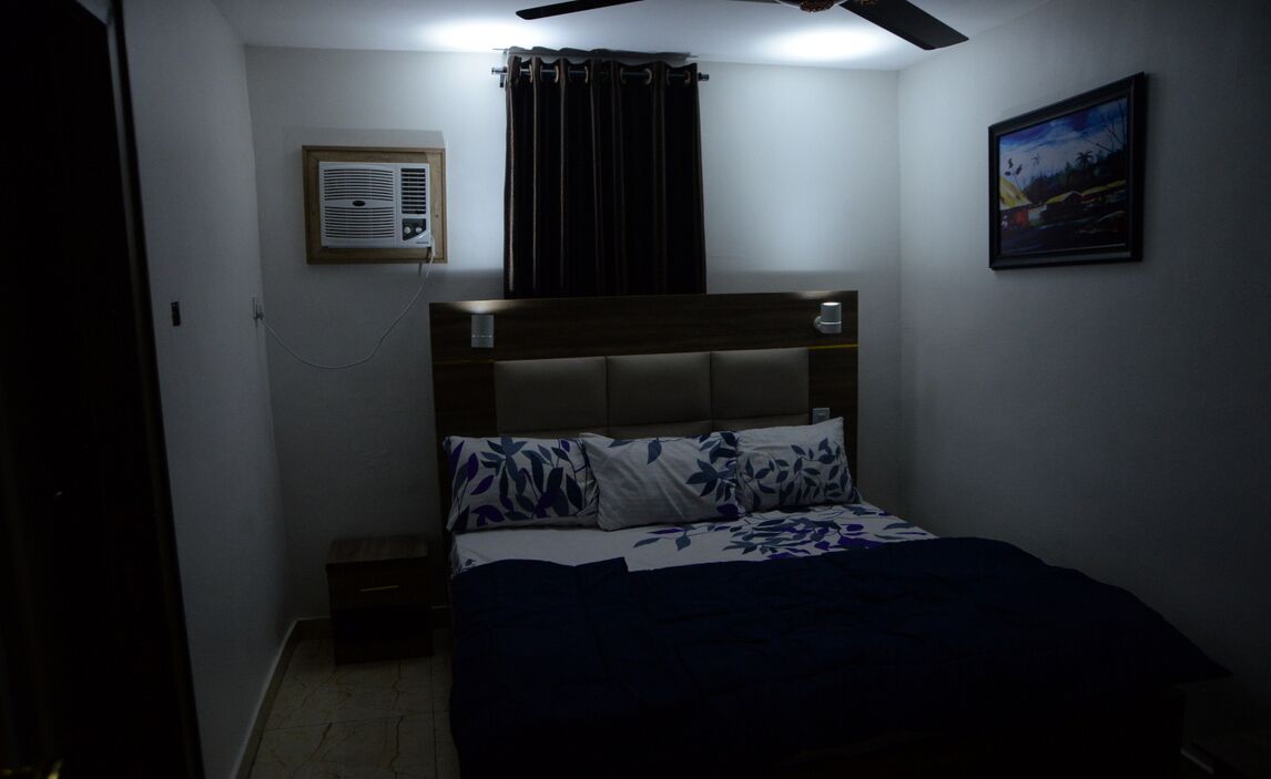 A bedroom with a bed, a ceiling fan, a wall-mounted air conditioner, a window with curtains, and a framed picture on the wall.