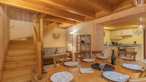 A wooden dining area with a round table set for six, a staircase, and a kitchen in the background. - LA Piemontaise - LE Penatsot (AUSSOIS)