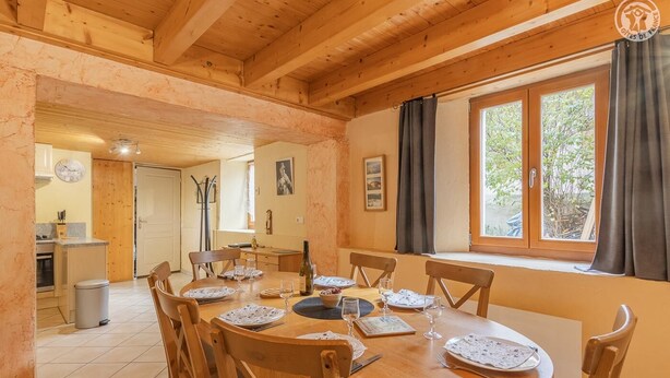 A wooden dining room with a round table set for four, chairs, and a window with curtains. - LA Piemontaise - LE Penatsot (AUSSOIS)