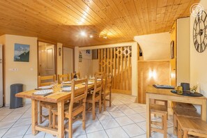 A wooden dining area with a long table and chairs, a clock on the wall, and a door with a wooden gate. - LA Piemontaise - LE Tsozal (AUSSOIS)