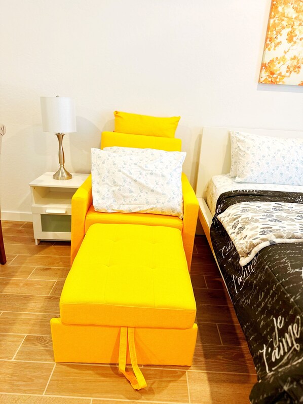 Una cama con cabecera y piecera amarillos, una mesita de noche blanca con una lámpara, y una pared con un cuadro floral.