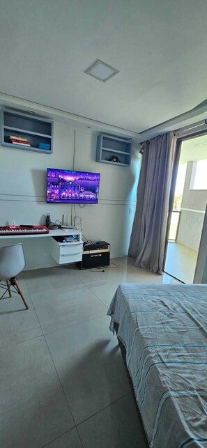 Ein Schlafzimmer mit einem Bett, einem Schreibtisch inklusive Tastatur, einem Fernseher und einem Fenster mit Vorhängen.