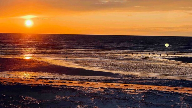 A beach at sunset with the sun low on the horizon, reflecting on the water.