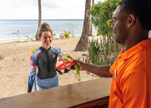 A person holding a water bottle and a towel on a sandy beach with palm trees and the ocean in the background. - Mai Sunset Beach Resort (Naviti Island)