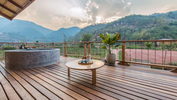 Une terrasse en bois agrémentée d’un bac à fleurs en pierre, d’une petite table décorée de divers objets, et offrant une vue sur un paysage montagneux.