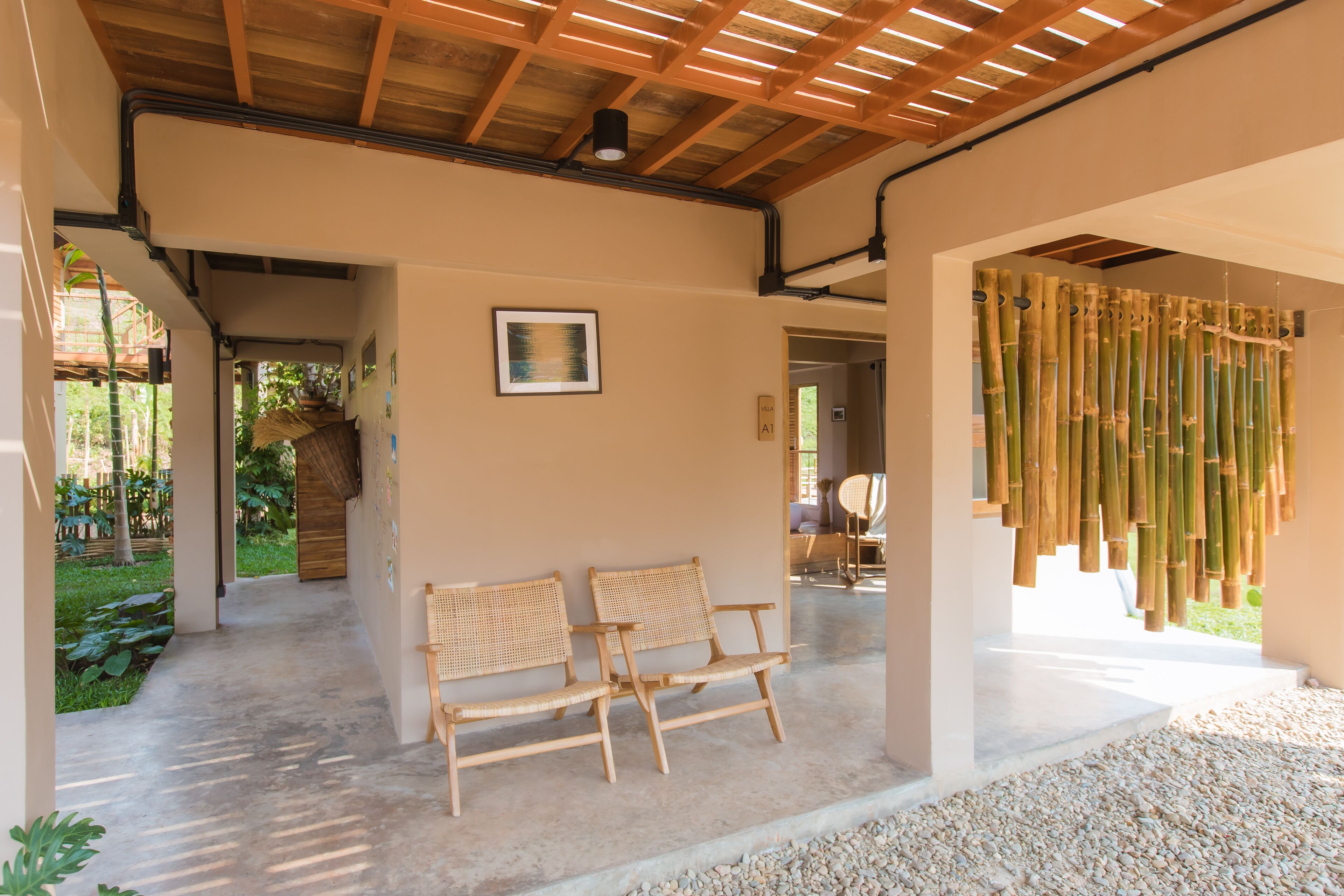 Un espacio interior con suelo de concreto, dos sillas de mimbre, techo de madera y cortina de bambú.